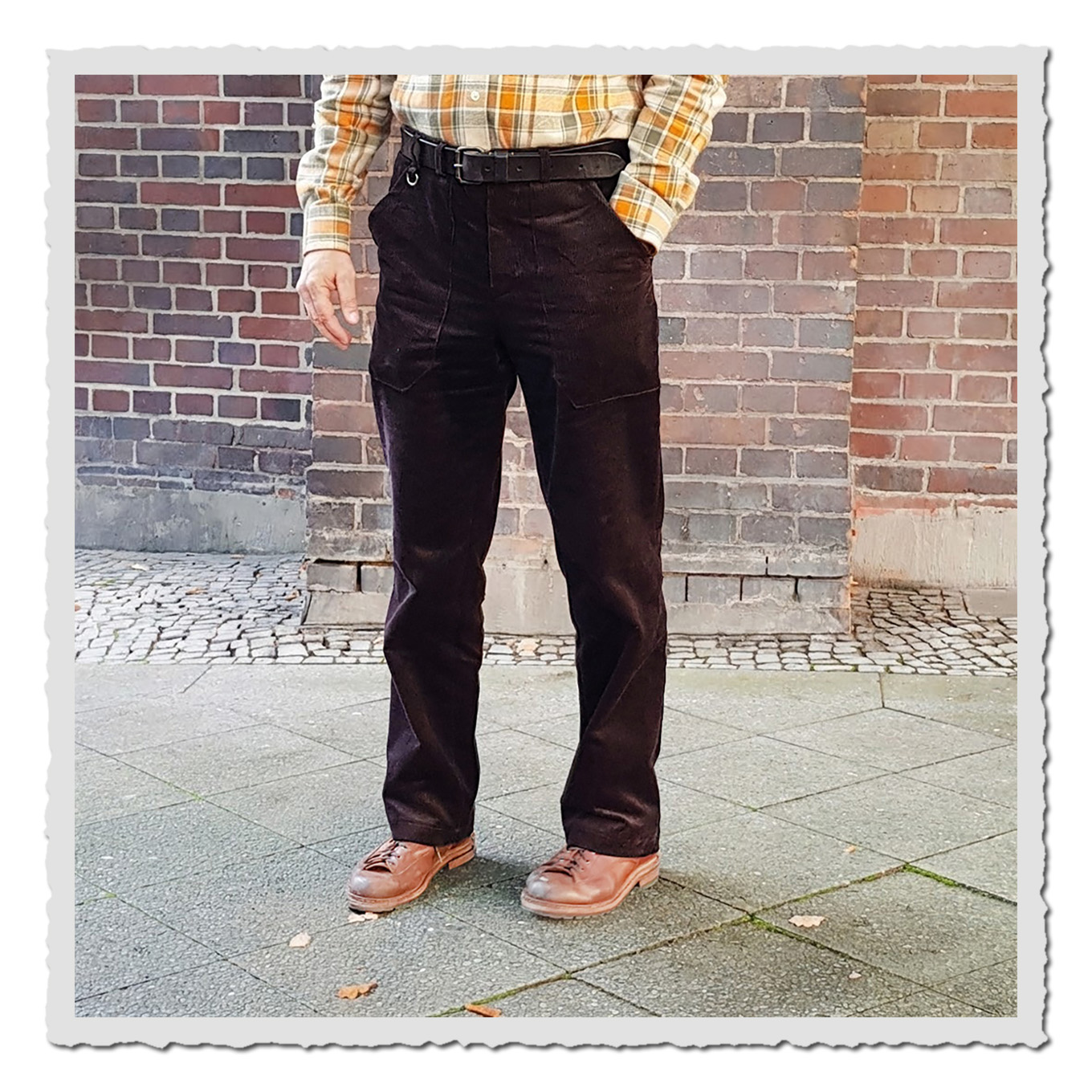 Person trägt schwarzbraune Cord-Arbeiterhose, Frontansicht vor Ziegelwand, Aufnahme draußen auf Gehweg.